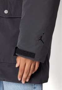 Svart vattentät jacka med en slät textur, med en Jordan-logotyp broderad på ärmen och en Velcro-stängning vid handleden.