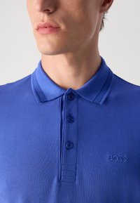Plava polo majica s teksturiranim materijalom, s tri gumba i izvezenim logom na lijevoj strani prsnog koša. U pratnji s rebrastim ovratnikom.