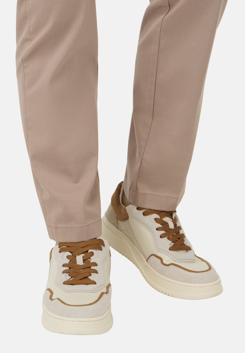 Béžové semišové a kožené tenisky s hnědými akcenty, vybavené kulatými tkaničkami a silnou bílou podrážkou. Nosené s světlými béžovými kalhotami.