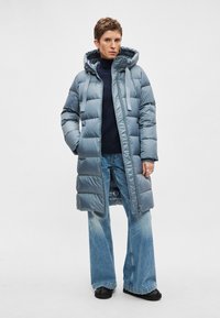 Gewatteerde blauwe donsjas met capuchon, voorzien van een ritssluiting en trekkoordbanden, gecombineerd met wijde, vervaagde jeans en zwarte schoenen.