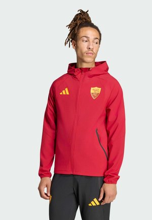 Uomo con dreadlocks che indossa una giacca rossa con cappuccio e pantaloni neri con loghi gialli, che guarda leggermente verso sinistra su uno sfondo uniforme.