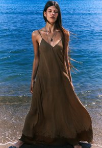Robe maxi marron fluide avec des bretelles spaghetti et une coupe décontractée, présentant une texture subtile et un ourlet légèrement plus foncé, portée par une personne se tenant près de l'eau.