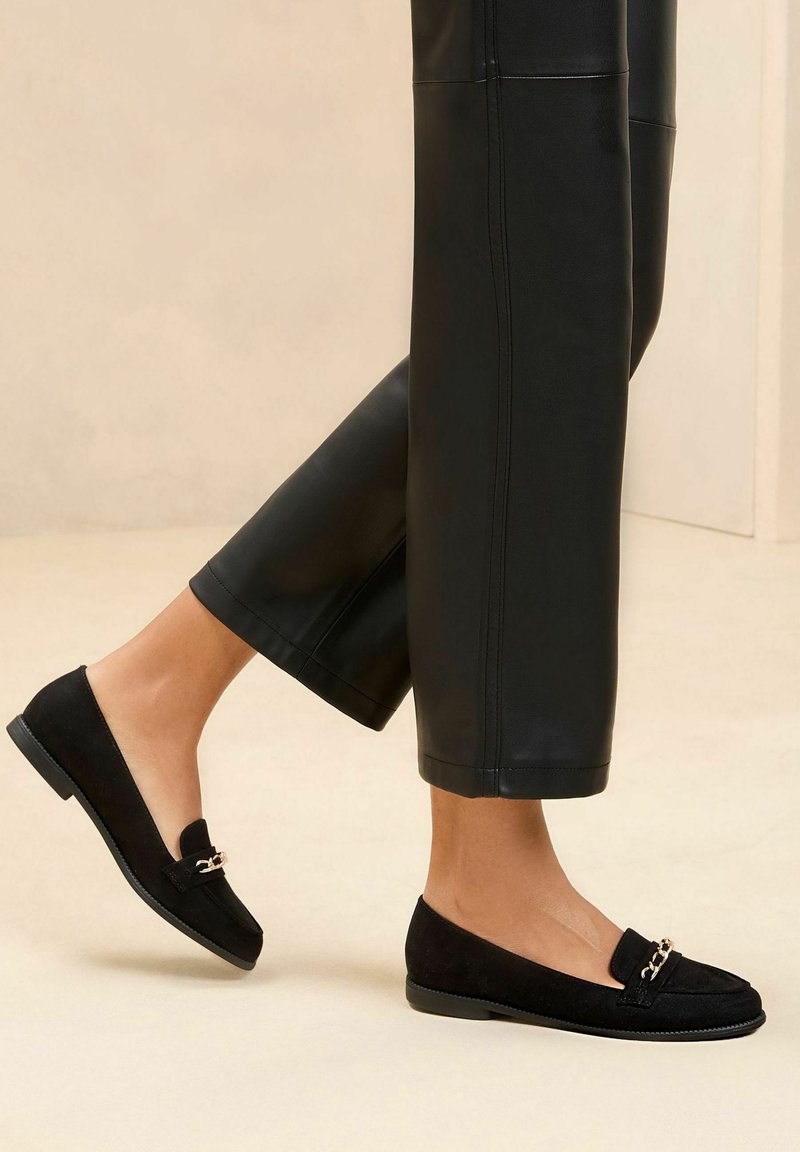 Black suede loafers with a flat sole, featuring a gold-tone chain detail on the front. Worn with black leather cropped pants.
