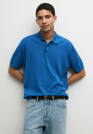 Junger Mann lehnt an einer weißen Wand, trägt ein leuchtend blaues Poloshirt, hellblaue Jeans und einen dunklen geflochtenen Gürtel, mit den Händen hinter dem Rücken.