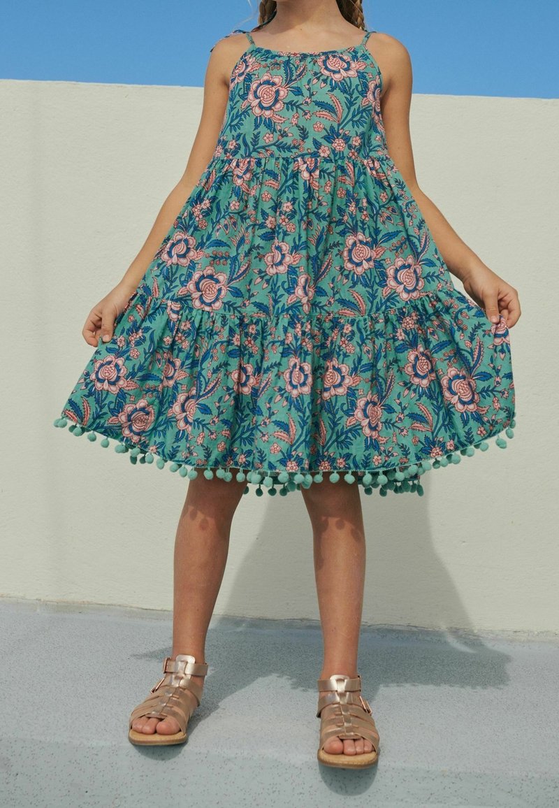 Enfant portant une robe sans manches turquoise avec motif floral rose et bordure à pompons, tenant les côtés, debout sur un sol gris contre un mur blanc.