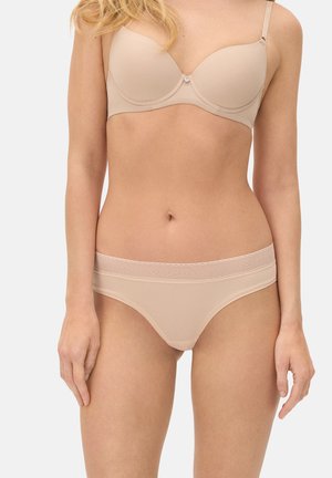 Mujer con un sujetador beige nude y ropa interior a juego con una cinturilla ancha y texturizada, posando frente a un fondo liso.