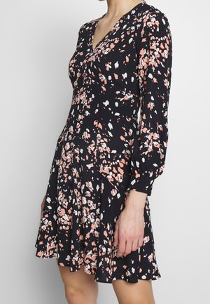 Kobieta ubrana w czarną sukienkę z różowo-białym abstrakcyjnym wzorem kwiatowym, z długimi bufkami na rękawach i rozkloszowaną spódnicą.