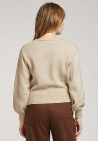 Pull en tricot beige avec un col rond, des poignets côtelés et une taille côtelée. Texture douce, avec des manches amples. Porté avec un pantalon marron.