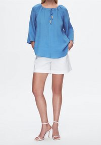 Blusa azzurra chiara con maniche a 3/4, scollatura con cordoncino e perline di legno; abbinata a pantaloni corti bianchi e sandali bianchi con cinturini.
