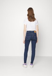 Vaqueros ajustados de denim azul oscuro con un parche de cuero marrón claro en la parte trasera y dos bolsillos traseros con detalles de costura. Combinados con una camiseta blanca.