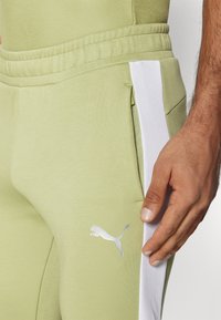 Pantalons de jogging vert clair avec des panneaux latéraux blancs, une taille élastique et un logo Puma argenté sur la cuisse gauche ; le tissu semble lisse et extensible.