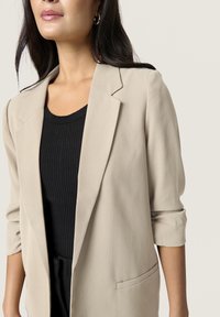 Blazer beige con spalle strutturate, maniche arrotolate e due tasche frontali, indossato sopra un top nero a coste. Tessuto dalla texture liscia.
