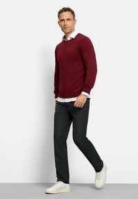 Bordeauxroter Pullover mit langen Ärmeln und Rundhalsausschnitt, über einem weißen Hemd mit Kragen getragen, kombiniert mit dunkler Hose und weißen Sneakers.