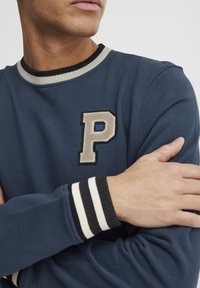 Navyblauwe sweatshirt met een geribde kraag, voorzien van een beige "P" applicatie en gestreepte zwart-witte manchetten. Zachte textuur is duidelijk zichtbaar.