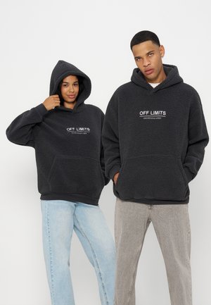 Två personer som bär svarta fleece-hoodies med texten "OFF LIMITS", kombinerade med ljusblå och beige byxor, stående mot vit bakgrund.
