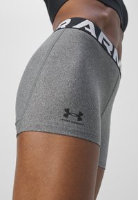 Gros plan d'une personne portant un short de compression gris Under Armour et un haut de sport noir sur un fond uni.