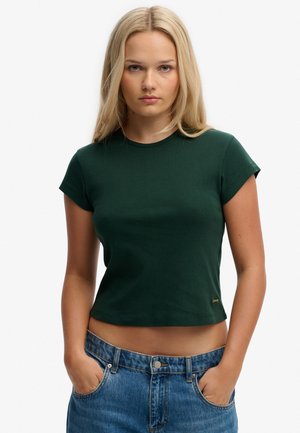 Mujer rubia con top corto de manga corta verde oscuro y vaqueros azules de tiro alto, con las manos en los bolsillos, mirando hacia adelante.