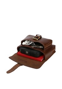Open brown leather bag containing white earphones, metal keys, black case, and red notebook on a white background.