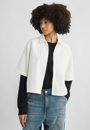 Γυναίκα με σγουρά μαλλιά, φορώντας λευκό μπουφάν με κοντά μανίκια και φερμουάρ πάνω από μαύρο πουκάμισο με μακριά μανίκια και μπλε τζιν, με τα χέρια στις τσέπες, κοιτάζοντας προς τα αριστερά.