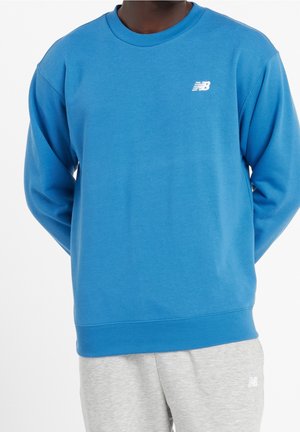 Mann trägt einen blauen New Balance Pullover und hellgraue Jogginghose, beide mit kleinen weißen New Balance Logos.