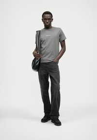 T-shirt cinza de algodão com logotipo branco, jeans de ganga pretos e calçado preto. O modelo segura uma bolsa preta de ombro, posando contra um fundo branco.