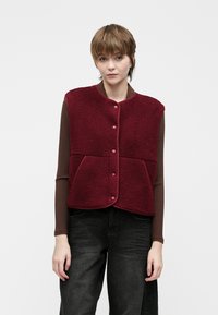 Mlada osoba u bordo flisu s dugim rukavima i dugim rukavima, s smeškom, nosi smeđu rebrastu majicu dugih rukava i tamne traperice, stoji naslonjena na jednostavnu pozadinu.