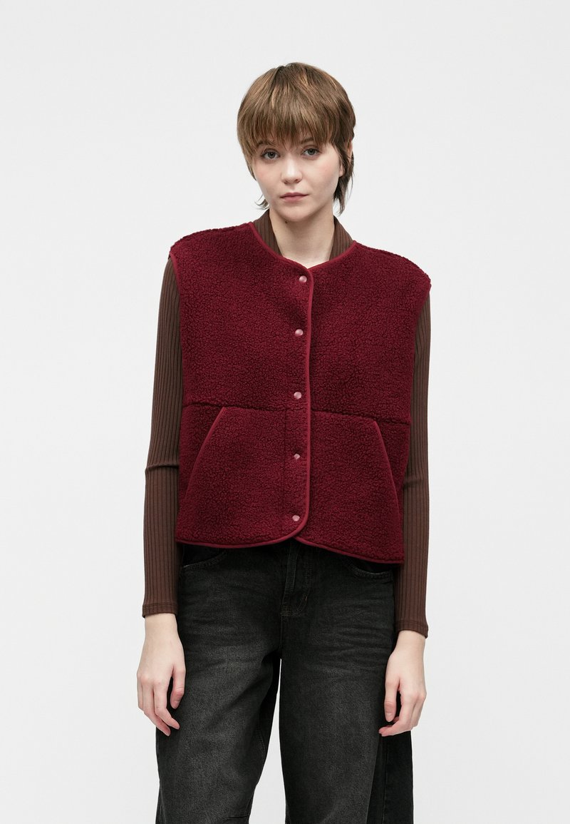 Mlada osoba u bordo flisu s dugim rukavima i dugim rukavima, s smeškom, nosi smeđu rebrastu majicu dugih rukava i tamne traperice, stoji naslonjena na jednostavnu pozadinu.