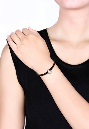 Un bracelet en cordon noir avec un fermoir circulaire en argent, porté au poignet, présentant un design lisse et minimaliste sur une chemise sombre.