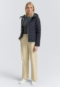 Giacca imbottita blu navy con un design trapuntato, abbinata a una maglietta a righe bianco e nera, pantaloni beige e sneakers bianche.