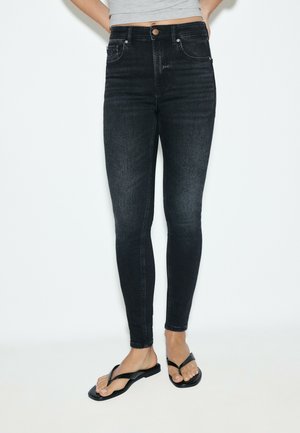 Personne portant un jean skinny noir taille haute avec fermeture par bouton, associé à des sandales plates noires et un top gris court.