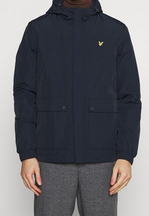 Mężczyzna ubrany w czarną kurtkę z kapturem, z klapkowymi kieszeniami i małym żółtym logo ptaka na piersi, w połączeniu z szaro-czarnymi spodniami w kratę.