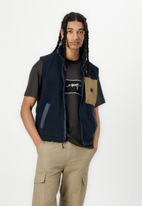 Mørkeblå fleecevest med en lys brun brystlomme, glidelås foran og sidelommer, brukt over en svart grafisk t-skjorte og beige cargo-bukser.