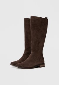 Botas altas castanhas em camurça, com uma textura suave, bico pontiagudo e um salto baixo em bloco. Apresentam um design simples e uma alça na parte de trás.