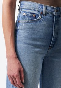 Vue de profil gauche d'une personne portant un jean en denim bleu clair taille haute avec un haut marron, la main reposant à côté de la cuisse.