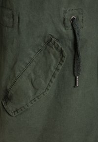 Ciemnozielony strój o teksturowanym wykończeniu. Posiada kieszeń naszywaną o kątownych krawędziach oraz sznurek z metalowym końcówkiem.