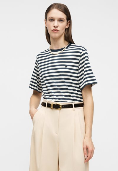 Γυναίκα που φοράει μπλούζα με ναυτικό και λευκό ριγέ, μπεζ παντελόνι ψηλόμεσο με μαύρη ζώνη και κολιέ με μαργαριτάρια, στέκεται μπροστά σε ένα απλό φόντο.