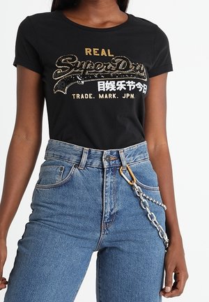Vrouw draagt een zwart SuperDry T-shirt met gouden en witte tekst en een high-waisted blauwe jeans met een zilveren kettingaccessoire aan de riemlus.