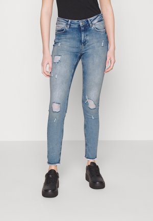 Femme portant un jean skinny bleu clair déchiré avec des ourlets effilochés et des baskets noires, debout devant un fond uni.