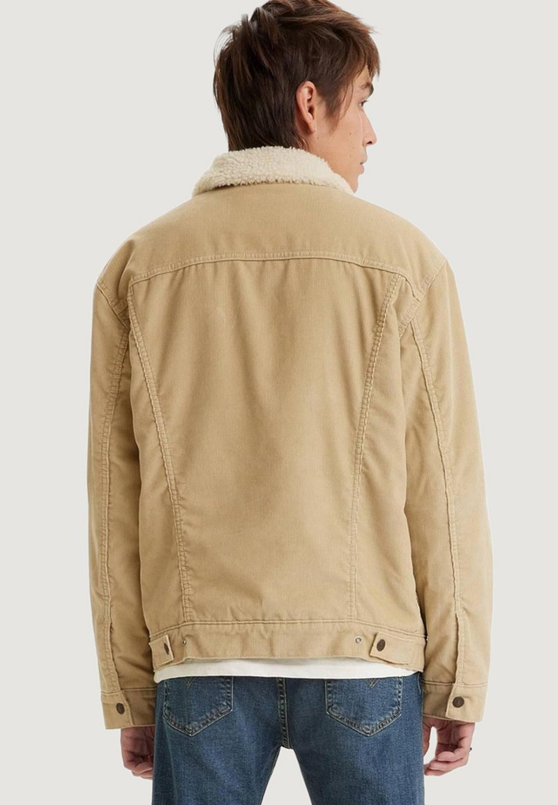 Chaqueta de pana beige con un cuello de piel sintética blanca, hombros estructurados y detalles de botones a presión en el dobladillo y los puños.