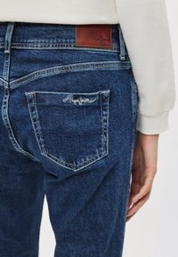 Jean en denim bleu avec poche arrière visible et un patch de marque en cuir marron au-dessus de la taille sur une personne portant un haut blanc.