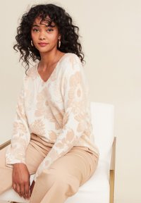 V-Ausschnitt Pullover in sanftem Creme mit pfirsichfarbener Blumenmusterung. Lässiger Schnitt mit langen Ärmeln, kombiniert mit hellbeigen Hosen. Schlichter Stuhl im Hintergrund.