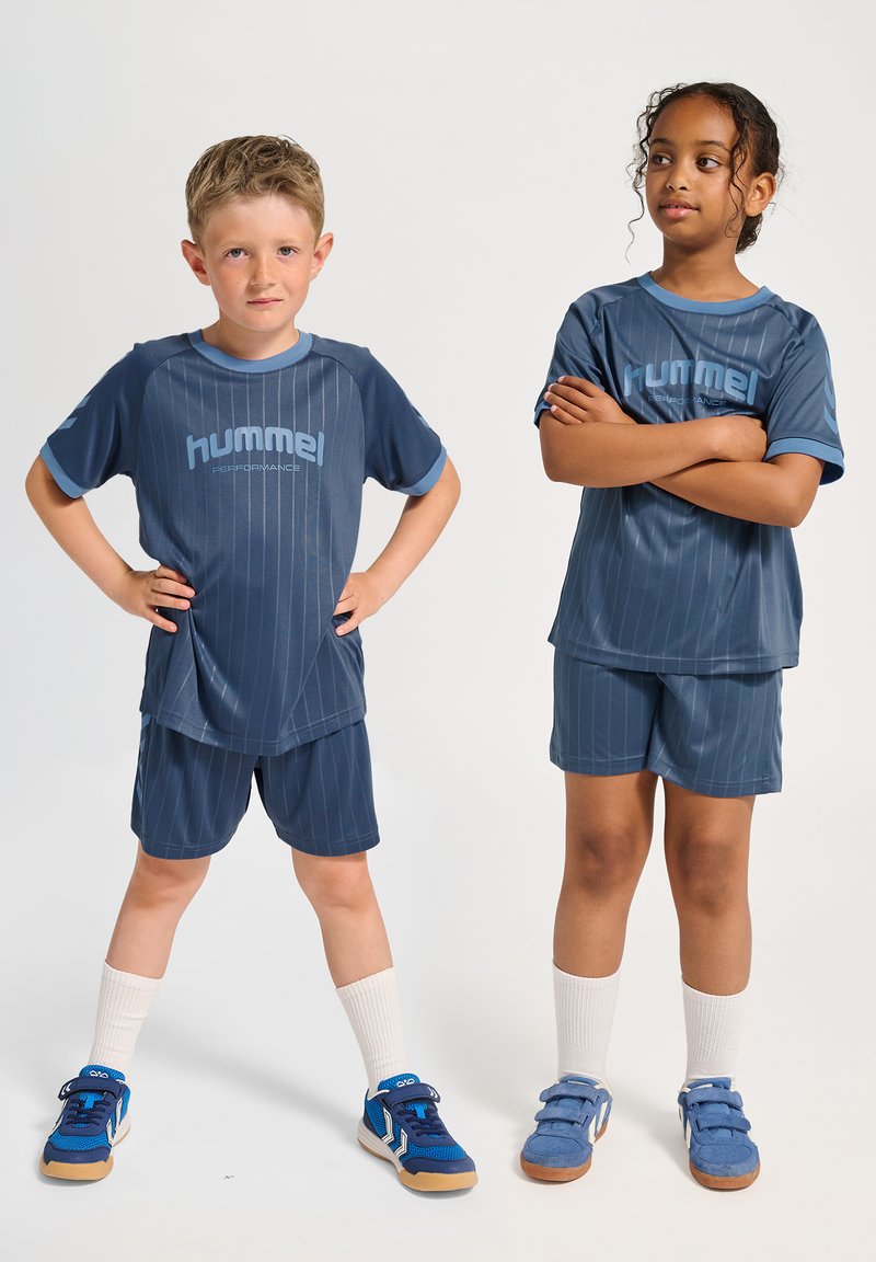 Niebieskie komplety sportowe z pionowymi paskami. Koszulki z krótkim rękawem i szorty, oba z napisem "hummel". Dopełnione sneakersami i białymi skarpetkami.