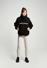 Fekete fleece dzseki magas gallérral, cipzáros záródással és kontrasztos fehér logókkal. Bézs leggingsgel és fekete sportcipővel viselve.
