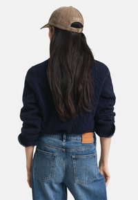 Granatowy sweter w warkocze z podwiniętymi rękawami, w połączeniu z niebieskimi dżinsami z skórzaną metką GANT z tyłu. Luźny styl na świeżym powietrzu.