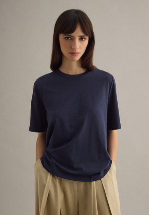 Donna con capelli castani lisci che indossa una maglietta a maniche corte blu navy e pantaloni beige plissettati, in piedi contro uno sfondo chiaro e uniforme.