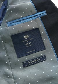 Mörkblå kostymjackas insida med ljusgrön foder som har små vita prickar. Etiketter för "NEXT" och "#LOOKSHARP" är synliga.
