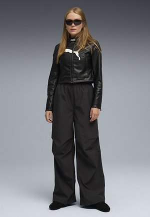 Chaqueta cropped de cuero negro con logo blanco, combinada con pantalones anchos negros. Los accesorios incluyen gafas de sol oscuras y un diseño elegante.