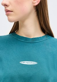 Νεαρός με μακριά καστανά μαλλιά που φοράει πετρόλ μπλούζα με λευκό λογότυπο "arkk copenhagen", εμφανίζεται από το λαιμό έως το στήθος με φόντο μονόχρωμο.