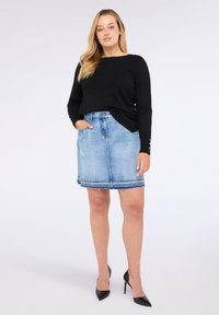 Vrouw staat met hand in de zak, draagt een zwarte lange mouwen top, een lichtblauwe spijkerrok met rafelrand en zwarte hoge hakken.