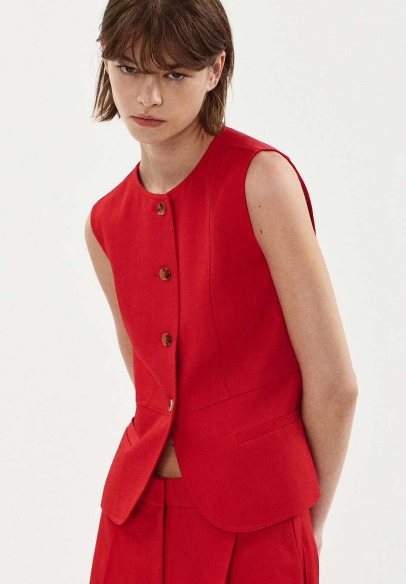 Jeune personne aux cheveux courts et bruns, portant un gilet sans manches rouge boutonné et un pantalon rouge assorti sur un fond uni.